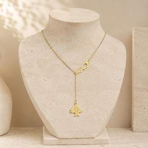 May include: A gold-toned lariat necklace featuring a delicate chain with two distinct charms. One charm is a small outline of the country of Lebanon, and the second charm is a detailed cutout of a cedar tree hanging from the centre of the chain.