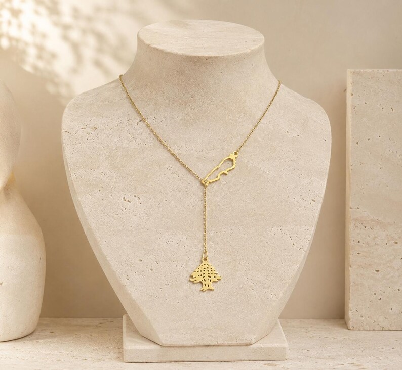 May include: A gold-toned lariat necklace featuring a delicate chain with two distinct charms. One charm is a small outline of the country of Lebanon, and the second charm is a detailed cutout of a cedar tree hanging from the centre of the chain.