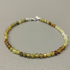 Pode incluir: Uma pulseira delicada com pequenas contas facetadas em tons de verde, castanho e amarelo. As contas são enfiadas com um fecho prateado. A pulseira é exibida em uma superfície cinza.