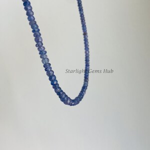 May include: A strand of blue gemstone beads, strung on a thin wire, hangs against a white background. The beads are small and round, with a slightly faceted appearance. The strand is long enough to be worn as a necklace.