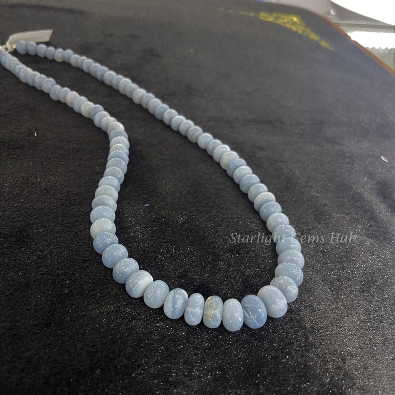 May include: A light blue gemstone necklace with round beads. The necklace is on a black surface with the text "Starlight Gems Hub".