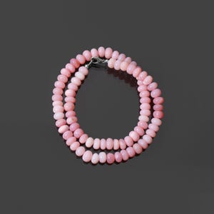 May include: A necklace composed of multiple strands of pink, oval-shaped beads. The beads are strung together, forming a continuous loop. A silver-colored clasp secures the necklace. The background is a dark, reflective surface.