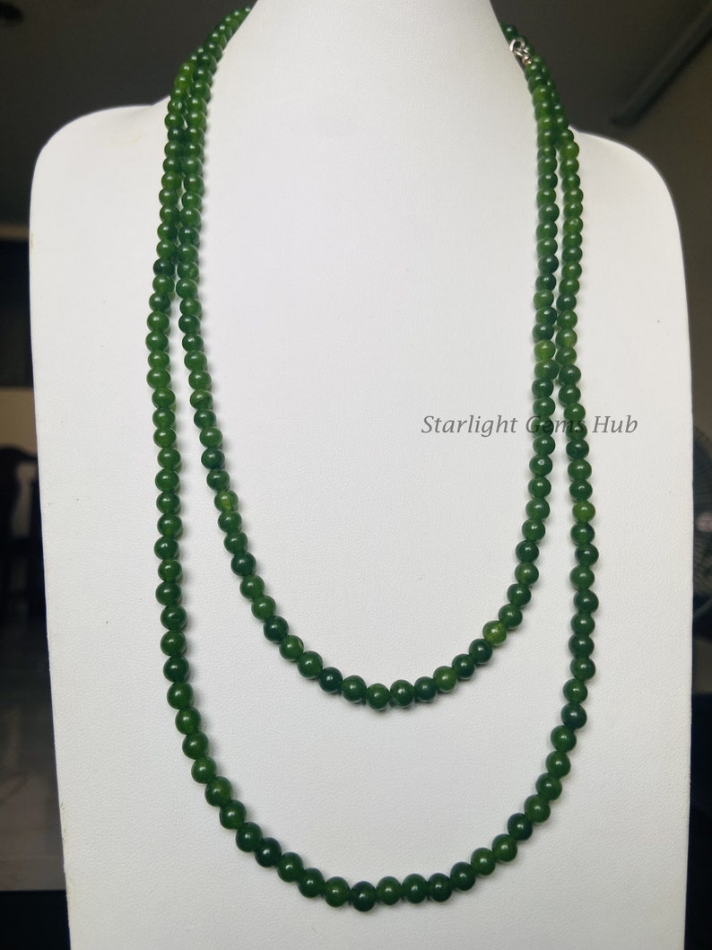 Puede incluir: Un collar largo de doble cadena hecho de cuentas de jade verde oscuro. El collar se muestra en un busto de maniqu&iacute; blanco.