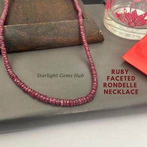 Könnte beinhalten: Eine Halskette aus roten Rubin-Edelsteinen, die aus facettierten Rondellen besteht. Die Halskette liegt auf einer grauen Oberfläche mit dem Text "Starlight Gems Hub" und "RUBY FACETED RONDELL NECKLACE".