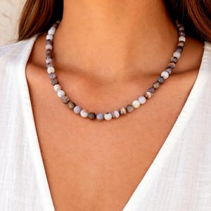 Op de afbeelding: Een ketting van kleine, ronde kralen in verschillende kleuren. De ketting heeft een mix van witte, bruine en grijze kralen. De ketting wordt gedragen op een witte top.