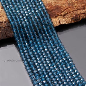 Puede incluir: Una hebra de cuentas de topacio azul, facetadas y de forma cuadrada, dispuestas en filas sobre una superficie de madera. Las cuentas son de color azul intenso y tienen un ligero brillo.  Starlight Gems está impreso en la superficie.