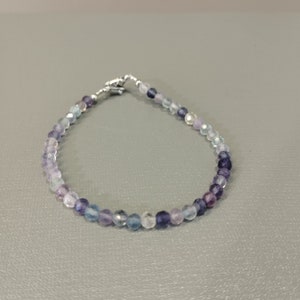 以下が含まれることがあります： 青、紫、透明な色合いの小さな丸い虹色のビーズで作られた繊細なブレスレット。ビーズはロブスタークラスプ付きのシルバーチェーンに糸で通されています。