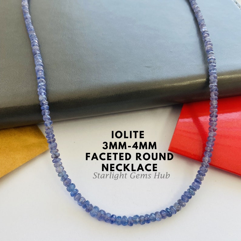 May include: A necklace made of faceted iolite beads. The beads are a light blue color and are approximately 3mm to 4mm in size. The necklace is on a gray surface and a red surface. The text "IOLITE 3MM-4MM FACETED ROUND NECKLACE Starlight Gems Hub" is visible in the image.