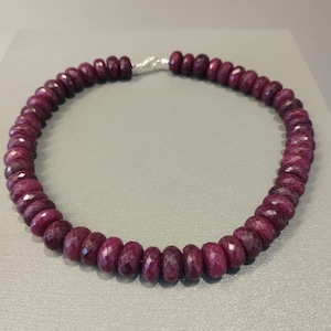 Op de afbeelding: Een ketting gemaakt van diep rode robijn kralen. De kralen zijn gefacetteerd en hebben een licht ovale vorm. De ketting is geregen op een zilveren ketting.
