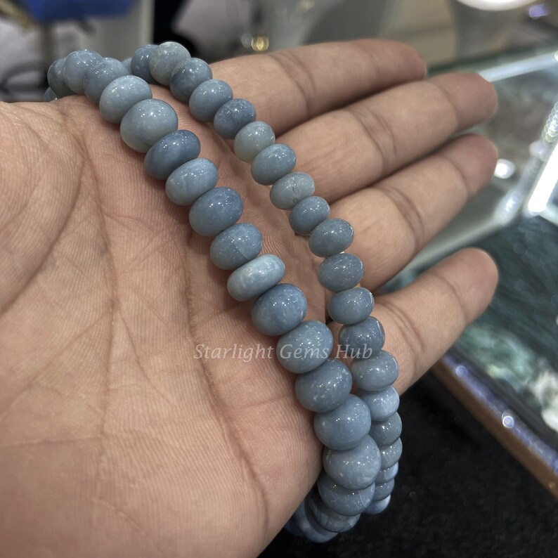 May include: A strand of light blue gemstone beads, likely opal, with a smooth, rounded shape. The beads are strung on a clear elastic cord and are approximately 8mm in diameter. The beads are displayed on a hand, with the text "Starlight Gems Hub" visible in the lower right corner.