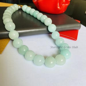May include: A light blue gemstone necklace with a silver clasp. The necklace is made of round beads and is laid out on a gray surface.