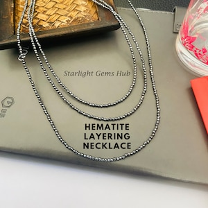 May include: Three layered necklaces made of small, dark gray hematite beads. The necklaces are displayed on a gray surface with the text "Starlight Gems Hub" and "HEMATITE LAYERING NECKLACE".