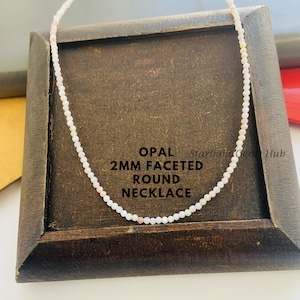 Puede incluir: Un collar de ópalo blanco hecho de cuentas redondas facetadas de 2 mm. El collar se muestra sobre una superficie de madera. El texto "OPAL 2MM FACETED ROUND NECKLACE" es visible en la superficie.