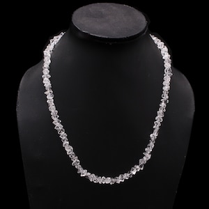 May include: A clear quartz crystal necklace on a black display stand. The necklace features small, irregularly shaped quartz crystals. The translucent crystals are strung together, catching the light and creating a shimmering effect.