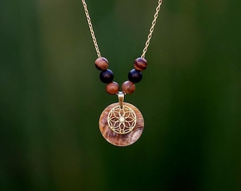 May include: A gold chain necklace featuring a round, iridescent shell pendant overlaid with a gold-toned geometric flower design. The necklace is accented with four dark brown and black wooden beads positioned above the pendant.