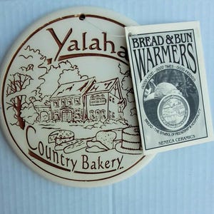 May include: A round, off-white ceramic bread warmer featuring a brown line drawing of the Yalaha Country Bakery building, trees, and baked goods. A rectangular paper tag is attached, reading BREAD & BUN WARMERS, GOOD FOOD, GOOD TIMES, GOOD FRIENDS.