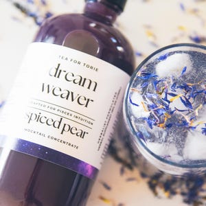 May include: A purple glass bottle with a white label that reads "Tea for Torie dream weaver crafted for Pisces intuition spiced pear mocktail concentrate". The bottle is sitting on a white surface with blue and yellow flower petals scattered around it. A glass with ice and blue flower petals is also visible.