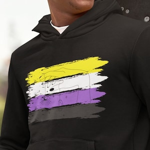 Puede incluir: Una sudadera con capucha negra con un diseño de rayas horizontales amarillas, blancas y moradas. Las rayas están pintadas en un estilo de pinceladas. El diseño es un símbolo de la identidad de género no binaria.