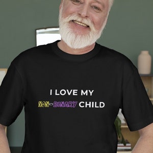 Puede incluir: Un hombre con una camiseta negra con el texto "Amo a mi hijo no binario" en blanco, con la palabra "no binario" en amarillo y morado. Está sonriendo y tiene barba blanca.