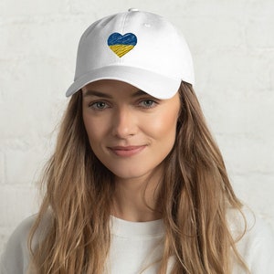 Könnte beinhalten: Weiße Baseballkappe mit einem blauen und gelben Herz, das auf der Vorderseite gestickt ist. Das Herz ist im Stil der ukrainischen Flagge.