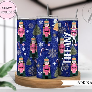 May include: Three stainless steel tumblers with a blue glitter background and a repeating pattern of nutcracker soldiers, Christmas trees, snowflakes, candy canes, and gold stars. The tumblers are on a wooden coaster. The text "TIFFANY" is on one of the tumblers. The text "STRAW INCLUDED" is in a pink circle at the top of the image.