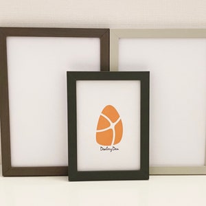 May include: Three picture frames, one brown, one grey, and one black, are arranged on a white surface. The black frame has a print of an orange abstract shape with the text "Dealery Den" underneath.
