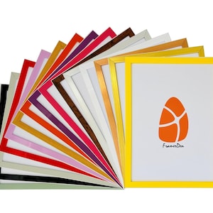 May include: A selection of picture frames in various colours, including green, black, white, pink, red, orange, yellow, gold, brown, purple, and blue. The frames are arranged in a fan shape, with the yellow frame at the front. The yellow frame features a small orange logo with the text "FramerDen" on it.