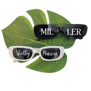 May include: Two pairs of sunglasses with personalized names. One pair is black with white text that says "MILLER". The other pair is white with black text that says "Shelby Marcus".