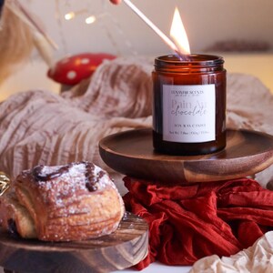 Może przedstawiać: Szklany słoik z brązowego szkła z białą etykietą z napisem "Luxe Home Scents Pain Au Chocolate Soy Wax Candle Net Wt. 7.2 oz (200g)". Świeca jest zapalona i płomień jest widoczny. Świeca stoi na drewnianej tacy.