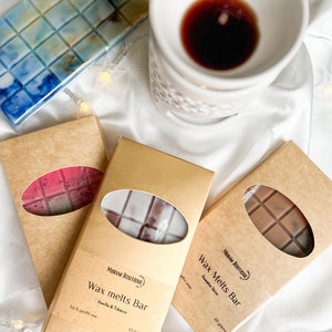 May include: An overhead shot features wax melt bars in various colors and scents, including blue, pink, and brown. The bars are in rectangular shapes and packaged in cardboard boxes. A white ceramic oil warmer with a dark liquid sits nearby.