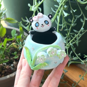 May include: A hand-painted, light blue panda planter with a smiling panda figure and a pink bow. The planter is decorated with green leaves, white flowers, and sparkling rhinestones. A charming decorative item for small plants.