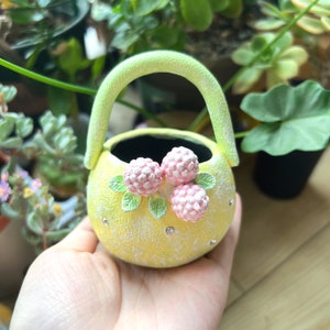 May include: A small yellow ceramic planter with a green handle and pink flower accents. The planter is decorated with white dots and has a black interior.