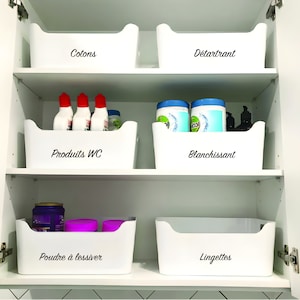 May include: White plastic storage bins labelled with black text in a white cabinet. The labels read "Cottons", "Detartant", "Produits WC", "Blanchissant", "Poudre à lessiver", and "Lingettes".