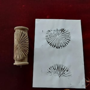 May include: A beige wooden roller stamp with a sunburst design, next to a white paper showing two stamped impressions of the same design in black ink. The stamp is cylindrical with a handle.