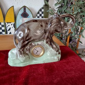 Puede incluir: Una figura de elefante de cerámica con un reloj. El elefante es marrón y gris, de pie sobre una base verde claro. El reloj tiene un marco dorado y una esfera blanca con números y manecillas negras.