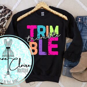 Può includere: Felpa nera con le parole "TRIM" in rosa, "trimble" in bianco, "BLE" in verde, blu e rosa. La felpa è su un appendiabiti e ha un aspetto usurato.