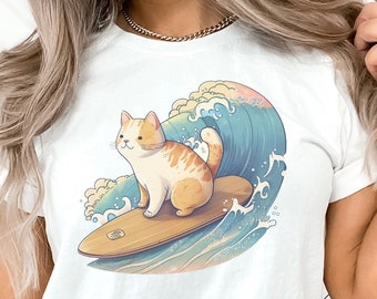 Puede incluir: Camiseta blanca con un gráfico de un gato surfeando en una ola. El gato es naranja y blanco y la ola es azul y verde.
