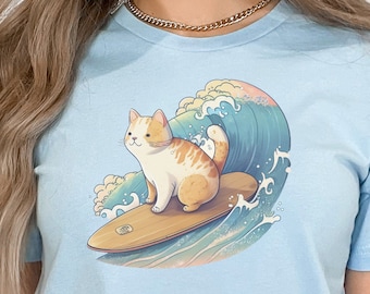Puede incluir: Una camiseta azul claro con un gráfico de un gato surfeando en una ola. El gato es naranja y blanco con una tabla de surf azul claro.
