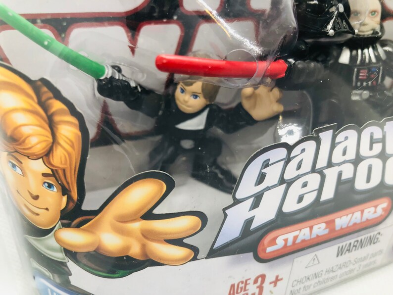 May include: A plastic package containing two Star Wars Galactic Heroes action figures. The figures are Luke Skywalker and Darth Vader. The package has a red, white, and black design with the words "Galactic Heroes" and "Star Wars" printed on it. The package also includes a warning label.