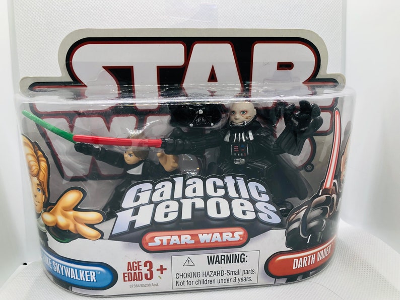 May include: A plastic package containing two Star Wars Galactic Heroes action figures. The figures are Darth Vader and Luke Skywalker. The package is red, black, and white with the Star Wars logo and the words "Galactic Heroes".