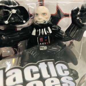 May include: A black and silver Darth Vader action figure with a white face and a black helmet. The figure is posed with one arm raised and the other arm bent at the elbow. The figure is in a plastic package with the words "Galactic Heroes" printed on the bottom.