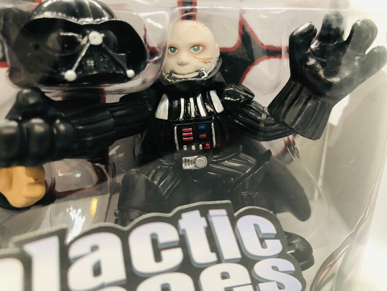 May include: A black and silver Darth Vader action figure with a white face and a black helmet. The figure is posed with one arm raised and the other arm bent at the elbow. The figure is in a plastic package with the words "Galactic Heroes" printed on the bottom.