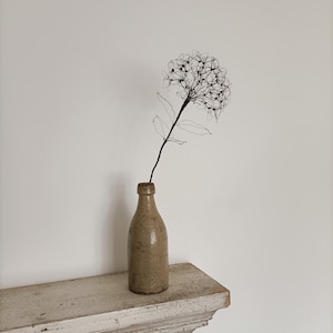 May include: A wire sculpture of a flower in a beige ceramic bottle on a white shelf.