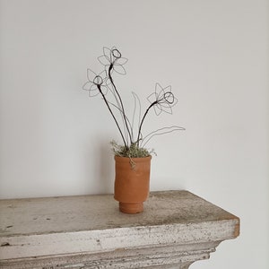 May include: A wire sculpture of a bunch of daffodils in a terracotta pot. The pot is sitting on a white shelf.