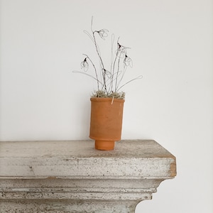 May include: A small terracotta pot containing a wire sculpture of delicate white flowers. The pot is sitting on a white shelf with a decorative edge.