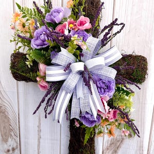 May include: A decorative cross made of woven twigs, adorned with purple, pink, and orange flowers, green foliage, and a large bow with gray and white stripes. The cross is set against a white wooden background.