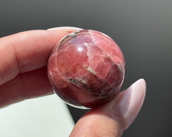 May include: A polished, spherical gemstone, predominantly pink with darker red and white veining. The crystal ball is held between fingers, showcasing its smooth, reflective surface. The gemstone is a natural mineral, likely used for decorative or spiritual purposes.