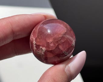 May include: A polished, spherical gemstone in shades of pink and red. The crystal ball is held between fingers, showcasing its smooth, reflective surface and natural color variations. The sphere is a decorative item.