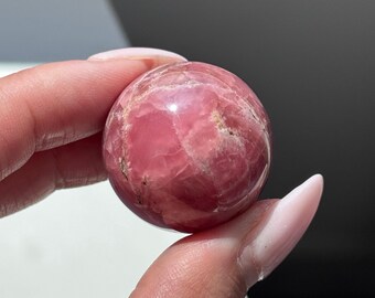 May include: A polished, spherical gemstone, primarily pink with lighter veining and subtle brown inclusions. The sphere is held between fingers, showcasing its smooth, reflective surface. The gemstone's color ranges from deep rose to pale pink.