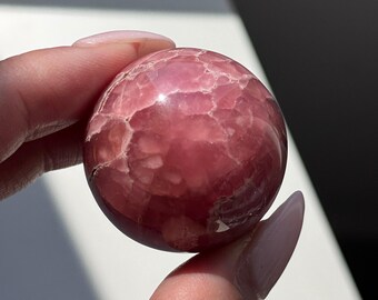 May include: A polished, spherical gemstone in shades of pink and white. The surface of the sphere has a cracked, textured appearance. The gemstone is held between two fingers.